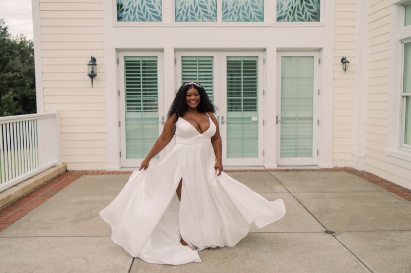 A smiling bride holding out the skirt of her fitted white gown on a patio
